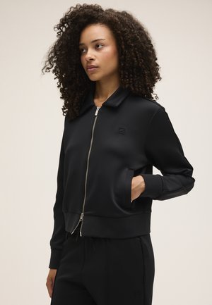 Frau mit lockigem Haar trägt eine schwarze Jacke mit Reißverschluss und passende Hose, steht mit einer Hand in der Jackentasche vor einem schlichten Hintergrund.