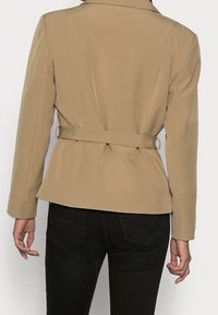 Béžová přizpůsobená bunda s páskem v pase, strukturovanými ramenními klouby a dlouhými rukávy. Hladká tkanina s čistým, minimalistickým designem.