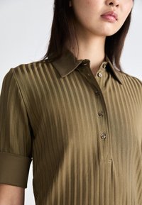 Olivenfarget, ribbestrikket polo skjorte med krage og korte ermer, med knappestengning foran og en subtil glans på stoffet.