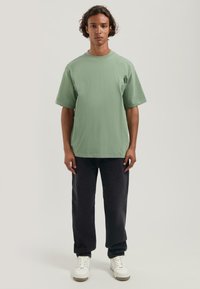 Camiseta de algodón verde con un corte relajado y mangas cortas, combinada con jeans oscuros y zapatillas blancas, resaltando un estilo casual.