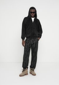 Μαύρο φούτερ με φερμουάρ και κουκούλα, με φαρδιά μανίκια και τσέπες μπροστά, συνδυασμένο με γκρι παντελόνι με σχέδια και ταμπά μποτάκια πεζοπορίας. Το μοντέλο φοράει γυαλιά ηλίου.