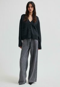 Sieviete, kas valkā brīvu melnu pogātu kardigānu ar kabatām un platajām izbalējušajām pelēkām džinsām, stāvot pret vienkāršu fonu.