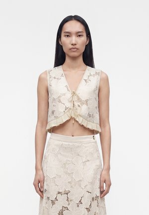 Žena s dlhými tmavými vlasmi, ktorá má na sebe bezrukávový béžový čipkovaný crop top s kvetinovými vzormi a zodpovedajúcu čipkovanú sukňu s vysokým pásom.