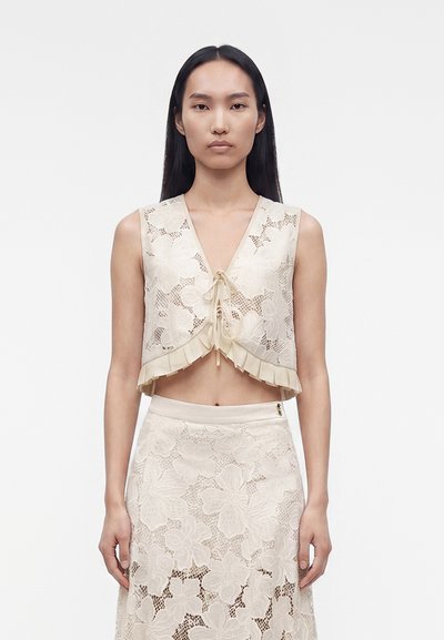 Femme aux longs cheveux foncés portant un crop top beige en dentelle sans manches avec des motifs floraux et une jupe en dentelle taille haute assortie.