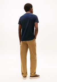 Mand stående med ryggen til kameraet, iført navyblå t-shirt, beige bukser med manchetter og brune sneakers mod en ensartet hvid baggrund.