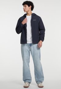 Giacca blu navy con dettagli in cucitura bianca, zip frontale e due tasche laterali, abbinata a jeans a gamba larga di un blu chiaro e sneakers beige.
