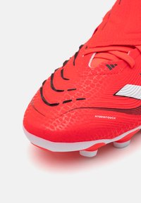 Red football boot with textured synthetic material, featuring black and white accents, a distinctive scale-like pattern, and a curved design.
