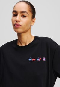 T-shirt en coton noir avec un graphique de quatre visages de dessins animés colorés portant des lunettes de soleil sur la zone de la poitrine. Design classique à col rond.