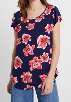 Kobieta nosząca granatowy top z krótkim rękawem z dużymi różowymi i czerwonymi kwiatowymi wzorami, połączony z jasnoniebieskimi dżinsami.