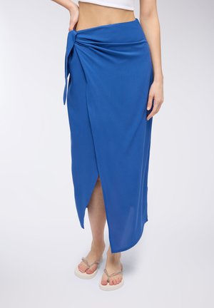 Gonna avvolgente blu realizzata in tessuto morbido, con dettaglio di nodo laterale e spacco alto, abbinata a ciabatte di colore chiaro.