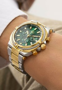 Gul och silver rostfritt stål klocka med en grön urtavla, flera undermått och kronograffunktioner. Texturerad bezel och tryckknappar.