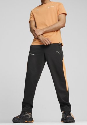 Pomarańczowa koszulka PUMA zestawiona z czarnymi dresami z pomarańczowymi akcentami. Gładki materiał, krótkie rękawy oraz detale z logo na obu elementach.