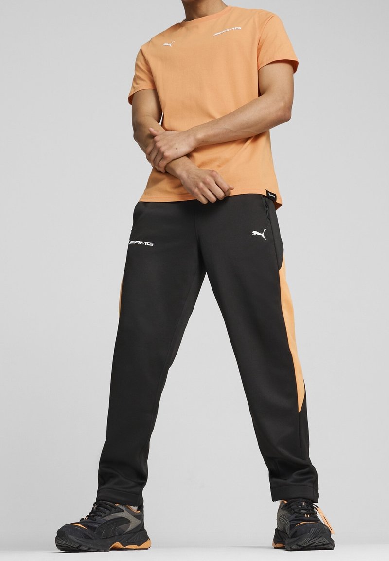 Pomarańczowa koszulka PUMA zestawiona z czarnymi dresami z pomarańczowymi akcentami. Gładki materiał, krótkie rękawy oraz detale z logo na obu elementach.