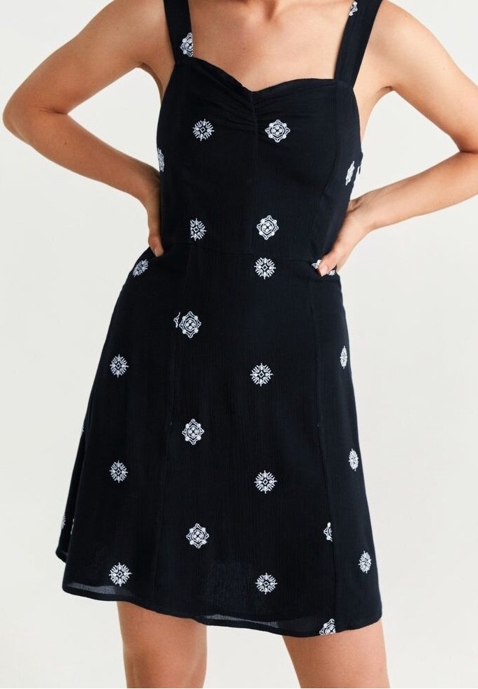 Robe noire avec encolure carrée, larges bretelles, et broderie florale blanche uniformément répartie. Tissu léger avec ourlet évasé.