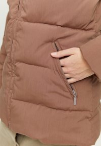 Brown puffer jacket with stitched sections and a zipped pocket on the side. Fabric appears smooth and slightly shiny.