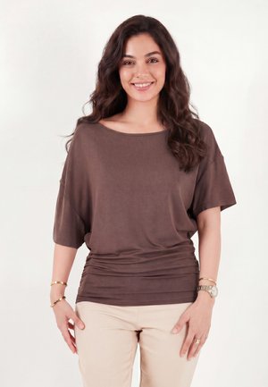 Jonge vrouw met lang golvend haar, die een losse bruine top, beige broek, gouden armbanden en een polshorloge draagt, glimlacht tegen een effen achtergrond.
