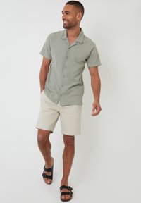 Camisa de botones de manga corta, color verde claro, con cuello camp. Combinada con pantalones cortos beige y sandalias negras. Textura suave, diseño casual.