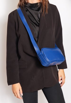Borsa a tracolla in pelle blu con forma rettangolare arrotondata, texture liscia e una larga tracolla regolabile. Indossata sopra un blazer nero.