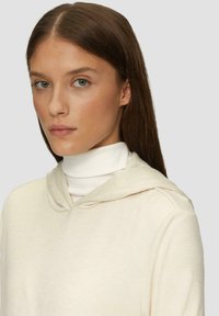 Crème-kleurige hoodie met een zachte textuur, voorzien van een trekkoordcapuchon en een gelaagde witte coltrui. Minimalistisch ontwerp met vloeiende lijnen.