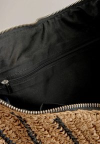 Vævet stråtaske med en textured naturlig tan overflade og sorte detaljer, som har en lynlåslukket sort stofforing og lynlåslukning.