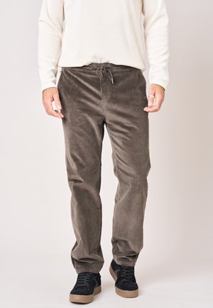 Man met grijze corduroy broek, wit overhemd met lange mouwen en zwarte sneakers met beige zolen, staand tegen een effen achtergrond.