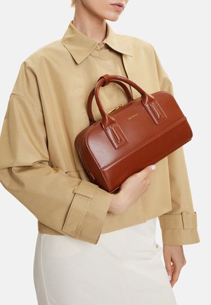 Donna con giacca beige che tiene una borsa di pelle marrone di media grandezza con doppi manici e dettaglio in cerniera dorata.