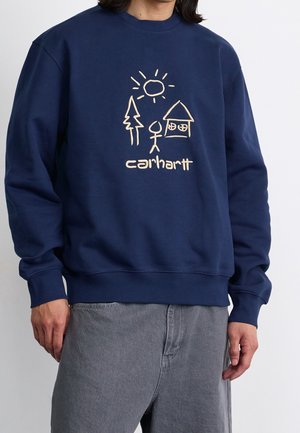 Mężczyzna w granatowej bluzie z kapturem z kremowym rysunkiem słońca, drzewa, ludzkiej sylwetki, domu oraz napisem „carhartt” na przodzie.