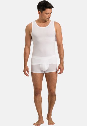 Man die blootsvoets staat en een nauwsluitend wit mouwloos ondershirt en witte boxershorts draagt tegen een effen achtergrond.