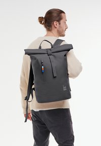 Sac à dos noir à rabat avec une poche zippée, présentant un matériau texturé gris et un accent coloré sur la tirette de la fermeture éclair. Logo affiché.