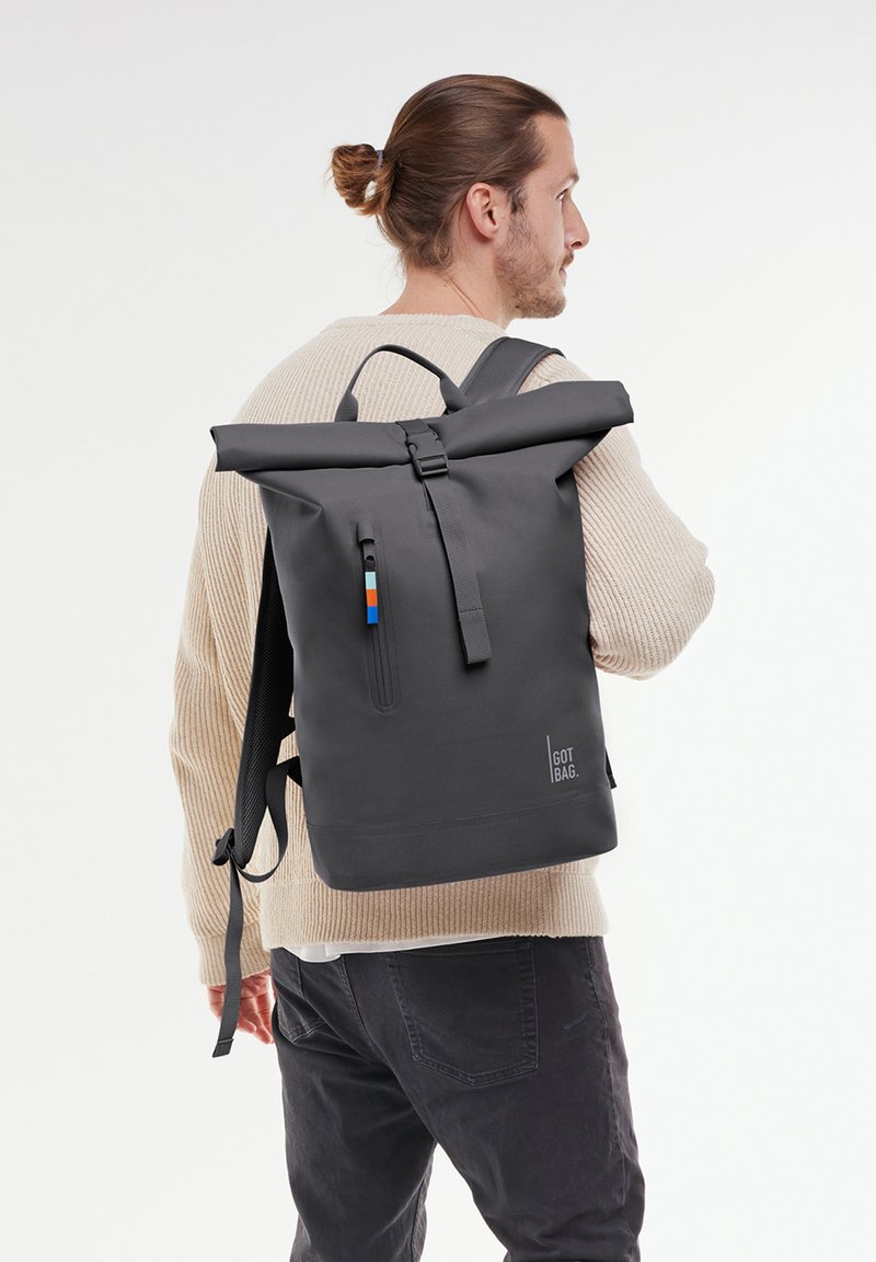 Sac à dos noir à rabat avec une poche zippée, présentant un matériau texturé gris et un accent coloré sur la tirette de la fermeture éclair. Logo affiché.