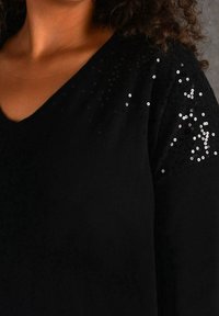 Schwarzer Pullover mit V-Ausschnitt und strukturiertem Stoff. Mit glänzenden schwarzen Pailletten an einer Schulter, die einen subtilen Akzent setzen.
