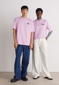 Due persone indossano T-shirt di cotone rosa, una delle quali ha una maglietta a maniche lunghe a righe bianche e nere sotto. I jeans blu e i pantaloni bianchi completano i top.