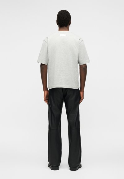 Άνδρας που στέκεται με την πλάτη, φοράει ανοιχτό γκρι κοντομάνικο πουκάμισο και φαρδιά μαύρη παντελόνια σε απλό λευκό φόντο.