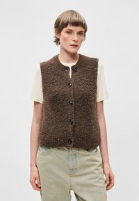 Colete castanho, texturizado, sem mangas, com um acabamento felpudo, apresentando um decote redondo e seis botões pretos na frente. Usado sobre uma camisa clara.