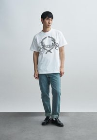 Λευκό T-shirt με κοντά μανίκια και μαύρο γραφικό σχέδιο που απεικονίζει δάφνινο στεφάνι και το κείμενο "Moschino", σε συνδυασμό με ανοιχτό μπλε τζιν παντελόνι και μαύρα παπούτσια.