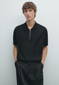 Černé polo tričko s kratkým zipem a strukturovanou tkaninou, krátké rukávy, kombinované s černými kalhotami na míru. Model stojí s rukama v kapsách před jednobarevným pozadím.