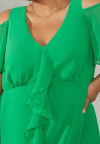 Grön klänning med V-ringning, flygande ärmar och en flödande volang längs framsidan. Tillverkad av lättviktsdukar med en jämn textur.