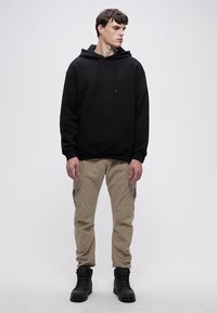 Crna majica s kapuljačom s prednjim džepom, uparena sa svijetlo bež cargo hlačama s bočnim džepovima i crnim čizmama sa vezicama.