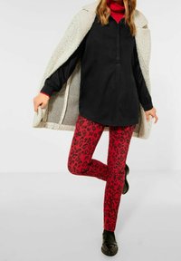 Zwarte blouse, rode turtleneck, rode legging met luipaardprint en zwarte schoenen. Een grijze textuurjas hangt over één schouder.