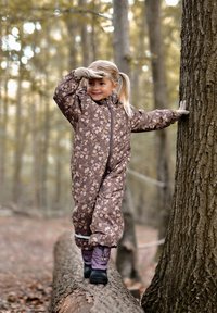 Salopette pour enfants à motifs floraux marron avec une fermeture éclair à l'avant, portée par-dessus un pantalon violet et des bottes noires, dans un environnement forestier.