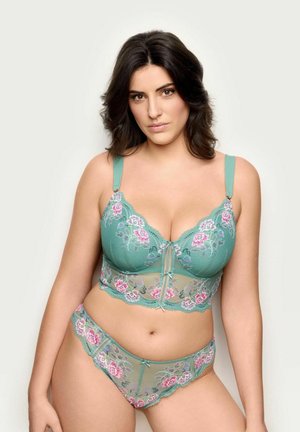 Mujer con lencería de encaje verde con bordado floral en rosa y blanco, posando contra un fondo liso y claro.