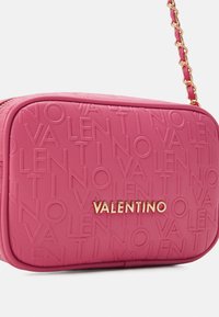 Sac en cuir rose embossé avec logo rép étitif, quincaillerie dorée, forme rectangulaire et bandoulière en chaîne. Détail de surface texturée visible.