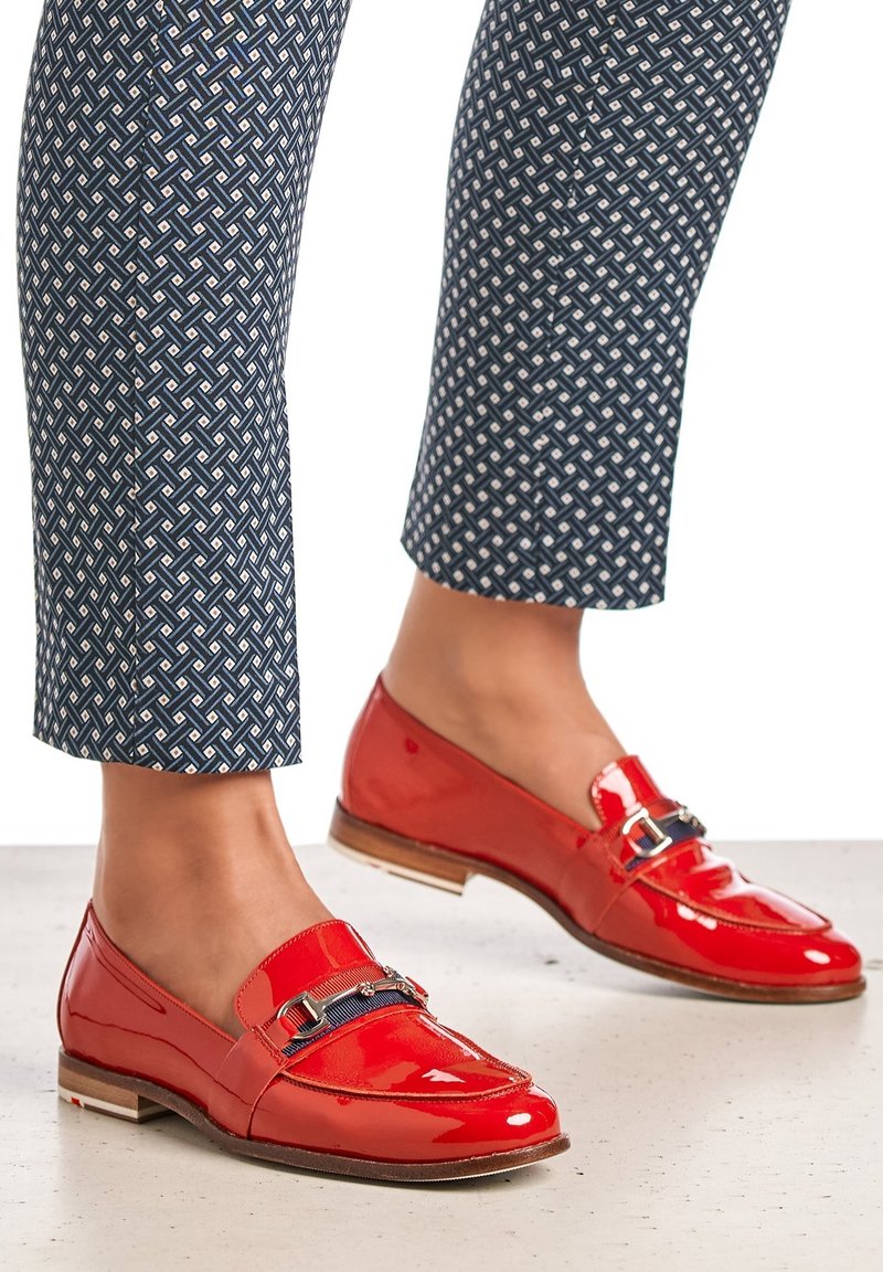 Pieds portant des mocassins rouges brillants avec des détails de boucle en métal foncé, assortis à un pantalon court bleu marine à motifs.