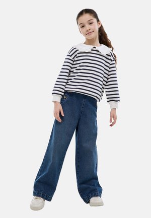 Fille aux longs cheveux attachés en queue de cheval haute, portant une blouse blanche à rayures noires avec un col en dentelle, un jean large bleu et des baskets blanches.