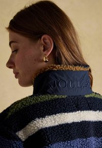 Fleece jacket with a navy and cream striped design, featuring a tan collar and embroidered 'JOULES' on the back. Gold hoop earrings visible.