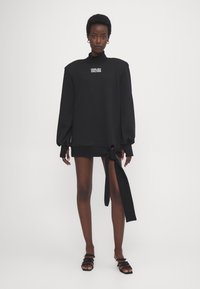 Sudadera negra de gran tamaño con mangas largas, cuello alto y logotipo frontal. Presenta un detalle de lazo en el dobladillo. Textura suave.