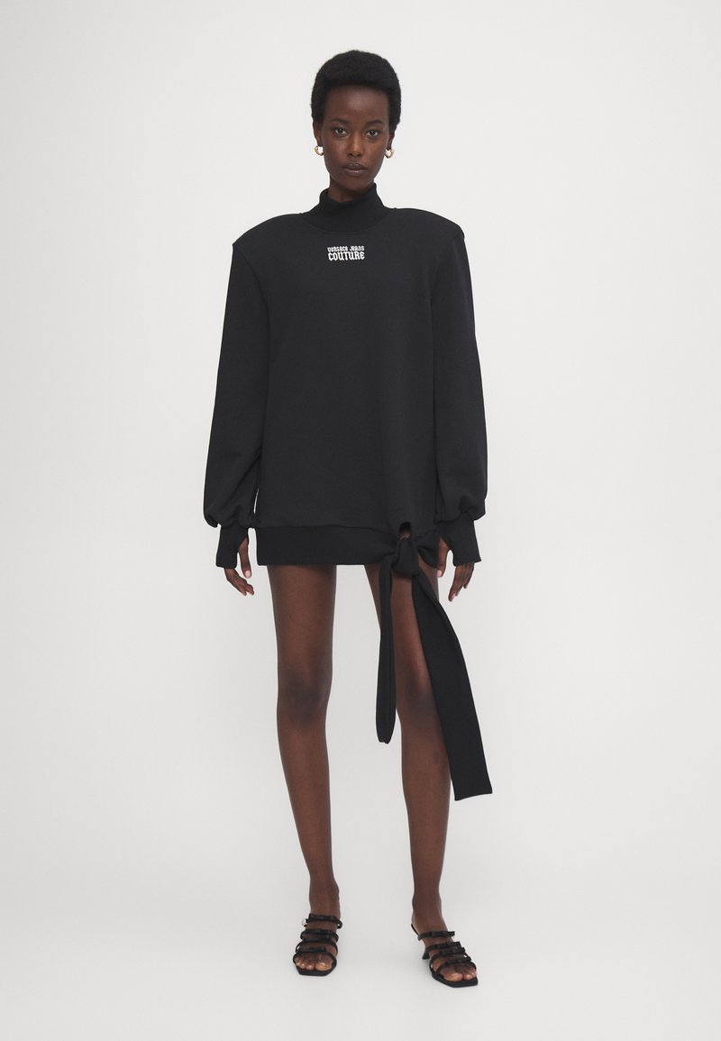 Sudadera negra de gran tamaño con mangas largas, cuello alto y logotipo frontal. Presenta un detalle de lazo en el dobladillo. Textura suave.