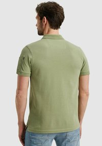 Grünes Polohemd mit kurzen Ärmeln und strukturiertem Kragen, seitlichem Taschen-Detail und glatter Textur, kombiniert mit hellblauen Jeans.