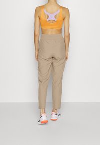 Kvinna som står vänd bort med en orange och lila sport-bh, beige högmidjade byxor och vita sneakers med orange sulor.