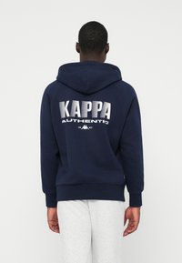 Moški obrnjeni proč, oblečen v temno modro kapuco s tekstom in logotipom "Kappa Authentic 1967" na hrbtni strani, v kombinaciji s svetlo sivo trenirko.